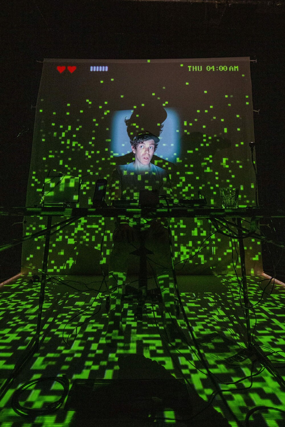 Tröll (photo credit: Tabitha Arthur) Image description: In a pitch black room, a young white man wearing a cap is visible from the shoulders up, illuminated by a square blue light shining up onto his face, with his enlarged silhouette visible behind him. Glitchy green lights are superimposed across the entire frame, and in the top left corner are two pixellated heart icons and several blue vertical bars, as if they were framing a vintage video game.