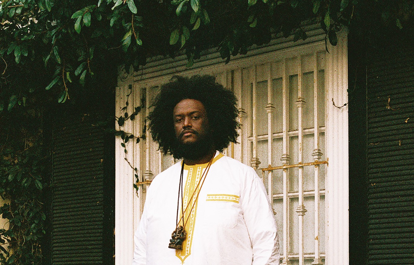 Kamasi Washington, a Black man dressed in a white shirt with a beard and voluminous hair, is pictured standing outside in front of a window. Green vines hang above the window.